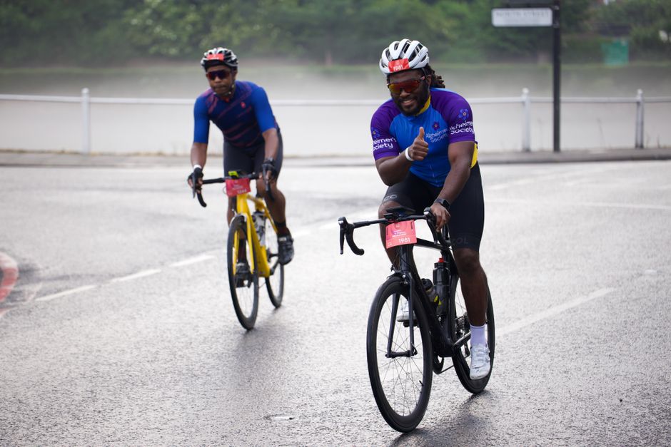 Two male cyclists riding on a paved outdoor path, wearing helmets and sports clothing. The man in the foreground is giving a thumbs-up gesture, smiling, and wearing a purple and blue cycling jersey, b