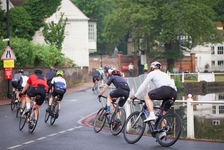 Two male cyclists riding on a paved outdoor path, wearing helmets and sports clothing. The man in the foreground is giving a thumbs-up gesture, smiling, and wearing a purple and blue cycling jersey, b